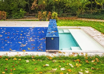 Une piscine partiellement couverte par une bâche bleue, entourée de pelouse verte et de feuilles d'automne dispersées.