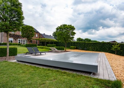 Terrasse avec piscine couverte grise, entourée de transats noirs, pelouse verte, arbres et haies, devant une maison en briques rouges.