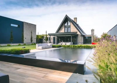 Maison moderne avec toit en pente entourée de jardin, piscine couverte au premier plan, et canapé extérieur gris clair sur la terrasse.