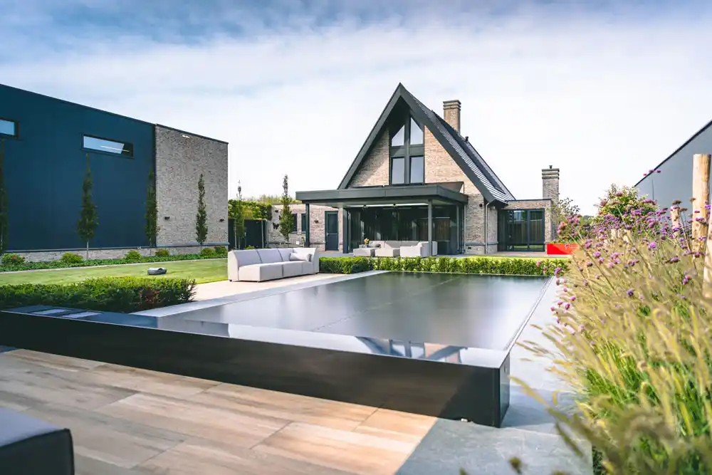 Maison moderne avec toit en pente entourée de jardin, piscine couverte au premier plan, et canapé extérieur gris clair sur la terrasse.
