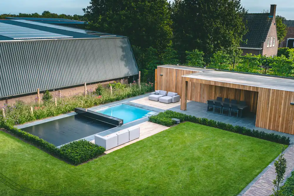 Une piscine avec couverture grise est entourée de mobilier de jardin et d'une terrasse en bois, avec pelouse verte et haies soignées.