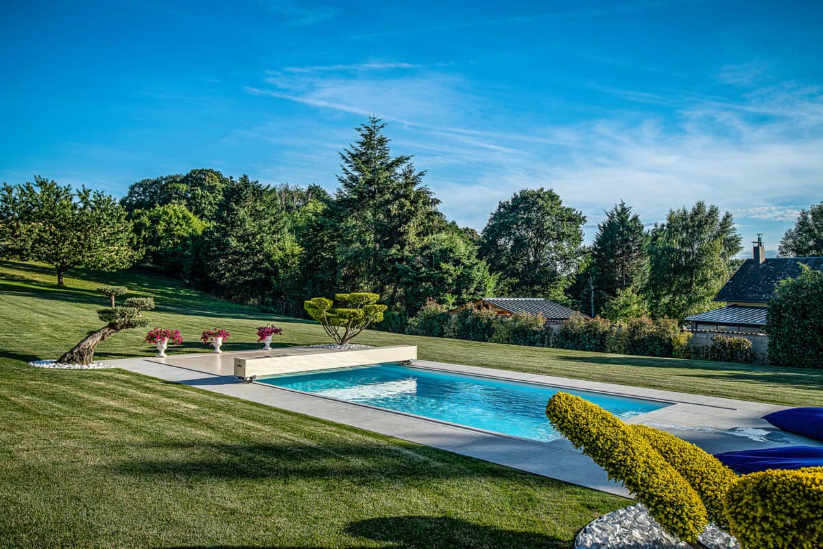 Jardin paysager avec piscine rectangulaire entourée de pelouse verte, arbres en arrière-plan et buissons taillés au premier plan.