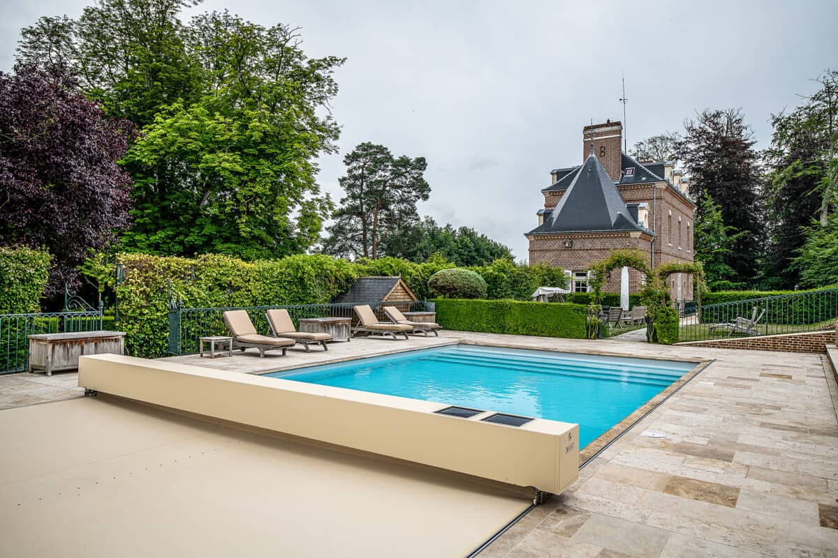 Piscine entourée de transats beige et d'une terrasse en pierre, avec un château en briques rouges à l'arrière-plan et des arbres verdoyants.