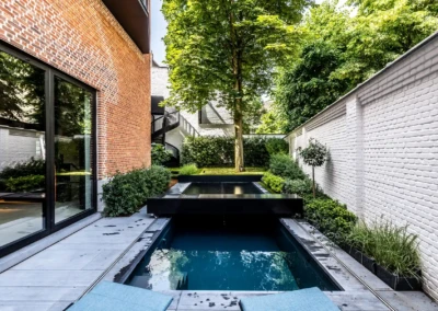 Chez-Anna-et-Luc-1 Une piscine rectangulaire entourée de murs en briques rouges et blanches, avec des plantes vertes et un arbre au fond.