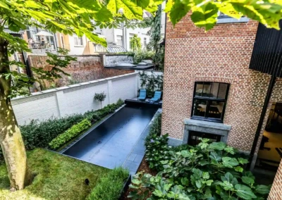 Chez-Anna-et-Luc-3 Cour arrière avec piscine couverte noire, entourée de murs en briques rouges et de végétation, deux chaises longues bleues visibles.