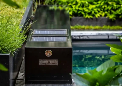 Chez-Anna-et-Luc-4 Boîtier noir avec panneaux solaires à côté d'une piscine, entouré de plantes vertes et de fleurs violettes.