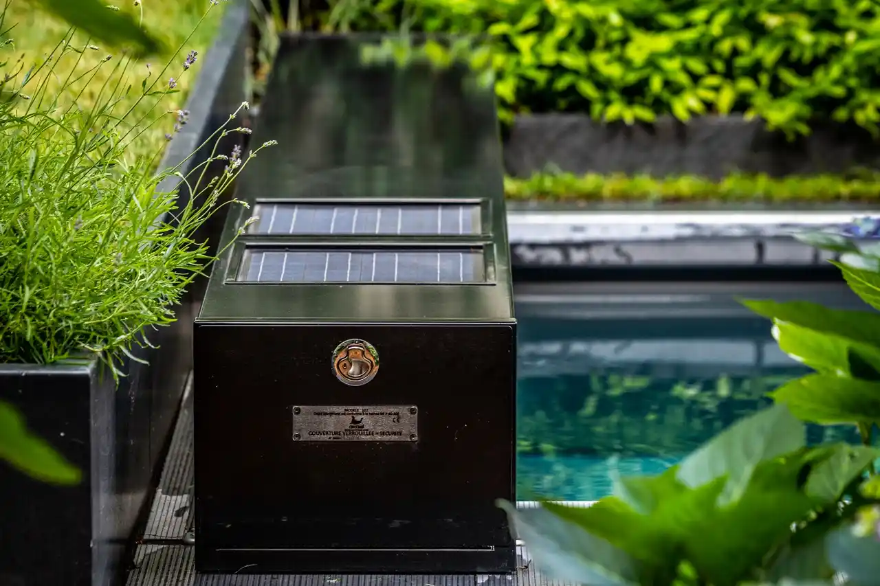 Boîtier noir avec panneaux solaires à côté d'une piscine, entouré de plantes vertes et de fleurs violettes.