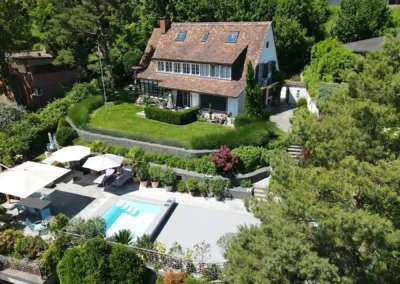 Maison avec toit en tuiles entourée de verdure, piscine et terrasse avec parasols blancs au premier plan.