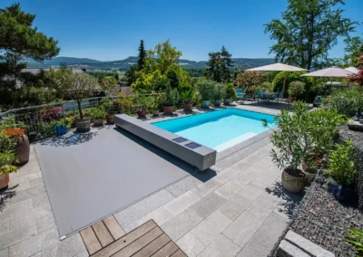 Piscine rectangulaire entourée de plantes en pot, avec une terrasse en dalles grises et parasols blancs sous un ciel bleu.