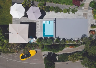 Vue aérienne d'une piscine rectangulaire entourée de parasols blancs et d'un jardin avec une voiture jaune garée à proximité.