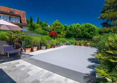 Terrasse avec piscine couverte de couleur grise, entourée de pots de fleurs et de verdure, sous un ciel bleu clair.