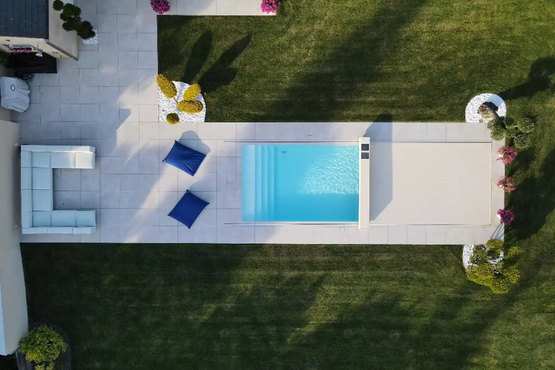 Vue aérienne d'une piscine rectangulaire, entourée de dalles blanches, avec des coussins bleus et des arbustes décoratifs sur pelouse verte.