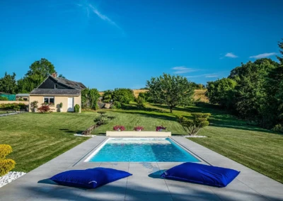 Chez-Gwendoline-et-François-3 Maison beige avec toit noir, piscine entourée de coussins bleus, pelouse verte et arbres en arrière-plan sous ciel bleu.