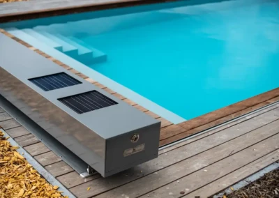 Une piscine avec une couverture grise dotée de panneaux solaires est entourée de margelles en bois et de copeaux de bois.