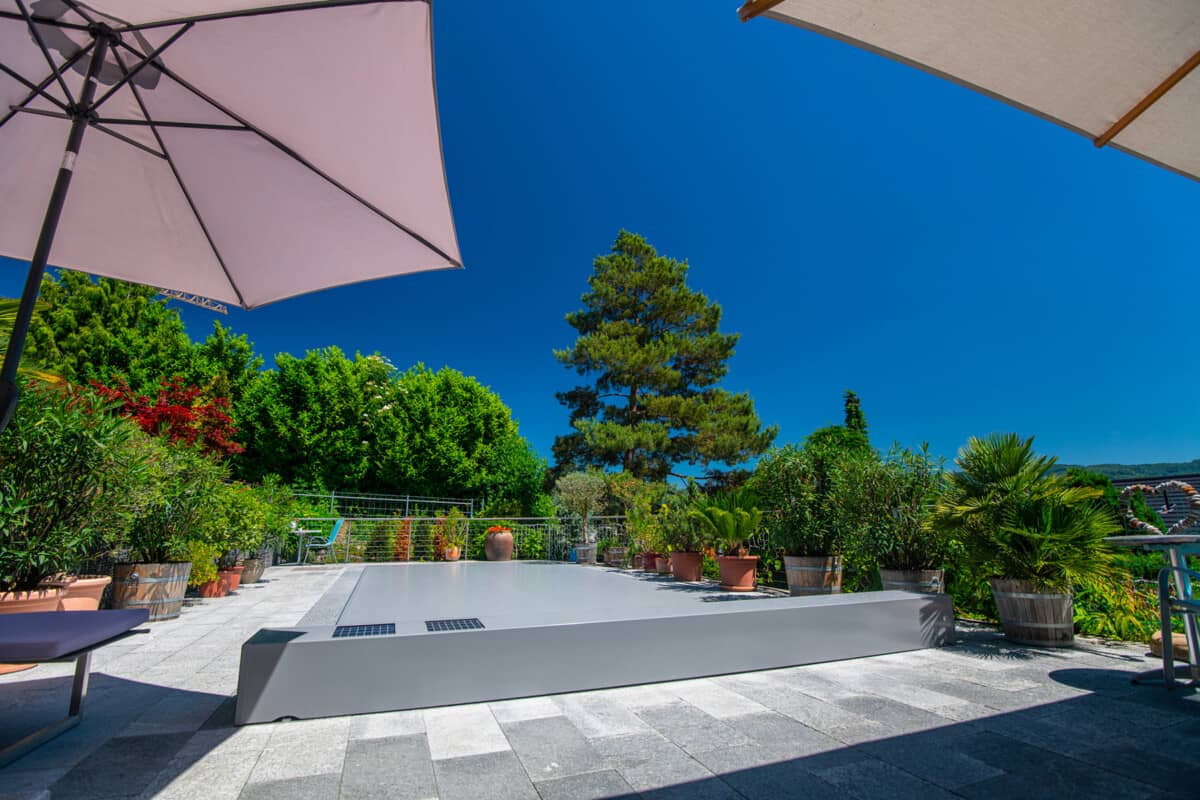 Une terrasse ensoleillée avec des parasols beiges, entourée de plantes en pots, et un arbre en arrière-plan sous un ciel bleu vif.