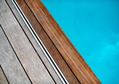 Planches de bois gris et brun clair bordent une piscine d'eau bleue, séparées par une fine barre blanche.