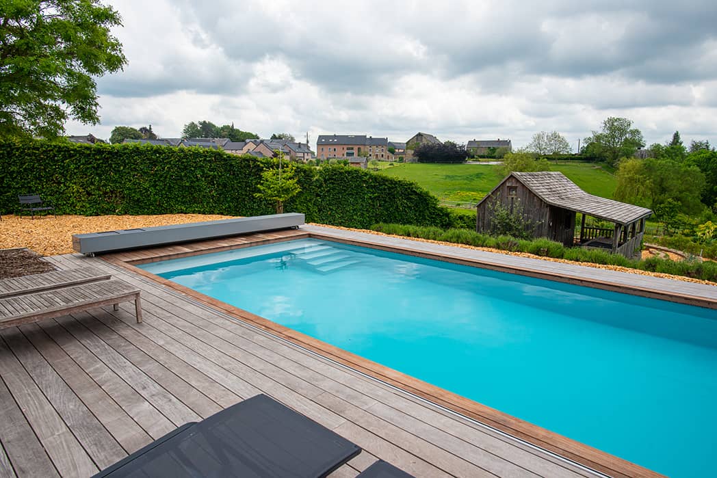 Piscine bleue entourée de terrasses en bois avec une haie verte en arrière-plan et des maisons à l'horizon sous un ciel nuageux.