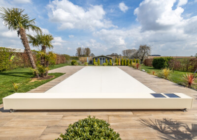 Terrasse en bois avec une couverture de piscine blanche, entourée de palmiers et d'arbustes, menant à un abri de jardin sous un ciel partiellement nuageux.