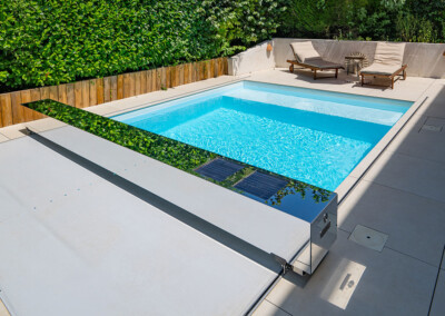 Vue large d’une piscine rectangulaire avec Coverseal semi-ouverte, terrasse claire et transats disposés à l’arrière, entourée de haies vertes.