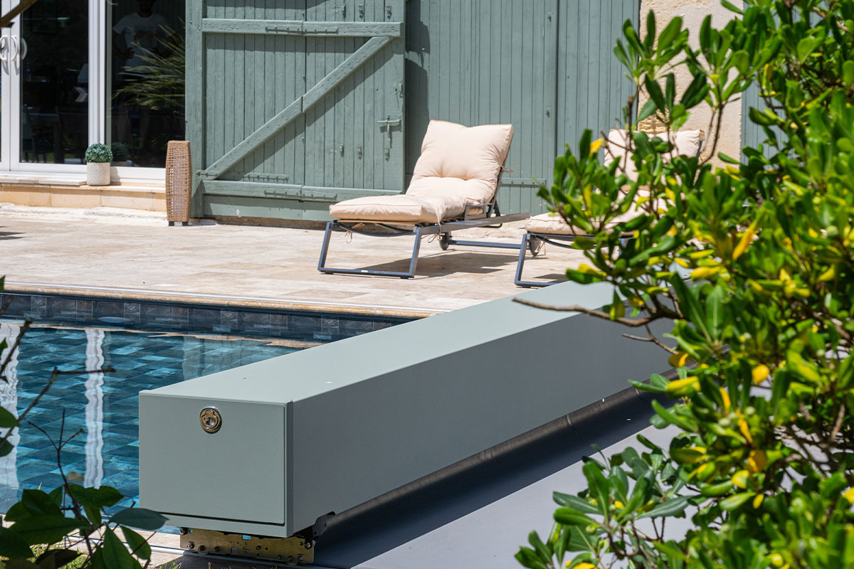 Gros plan sur l'habillage de la Coverseal grise au bord de la piscine, avec en arrière-plan les transats et la maison en pierre.
